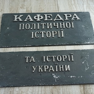 May include: Two dark grey rectangular metal signs with white lettering. The top sign reads "Кафедра Політичної Історії" and the bottom sign reads "Та Історії України".