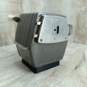 Peut inclure: Un taille-crayon vintage en métal gris avec une manivelle blanche. Le taille-crayon a un panneau en métal argenté avec un petit trou et le texte "BOSTON 500" sur le fond.
