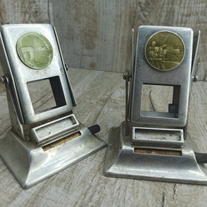 May include: Two vintage silver-toned metal stamp dispensers with a coin slot and a small window. One dispenser has a round image of a building, the other has a round image of people in uniform.