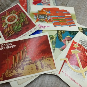 May include: A collection of vintage postcards with Soviet-era imagery. The postcards feature red and gold designs, including a star with a hammer and sickle, a battleship, and text in Cyrillic script. Some cards have military themes.