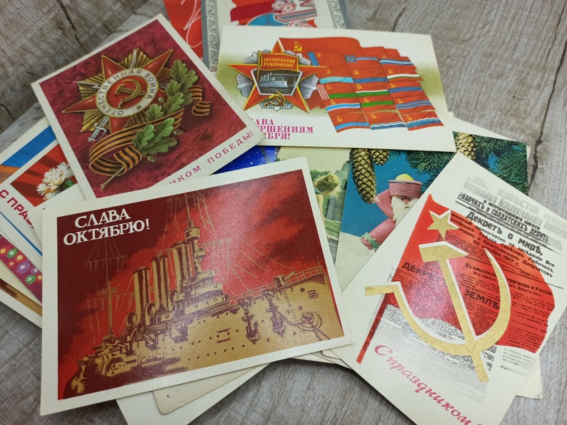 May include: A collection of vintage postcards with Soviet-era imagery. The postcards feature red and gold designs, including a star with a hammer and sickle, a battleship, and text in Cyrillic script. Some cards have military themes.