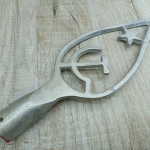 May include: A silver metal finial featuring a hammer and sickle and a star design. The finial is shaped like a leaf and has a hole at the bottom for attaching to a pole.