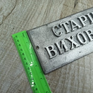 Puede incluir: Un letrero de metal plateado con el texto "СТАРШИ ВИХОВАТ" en letras en relieve. El letrero mide aproximadamente 15 cm de largo y 5 cm de ancho.
