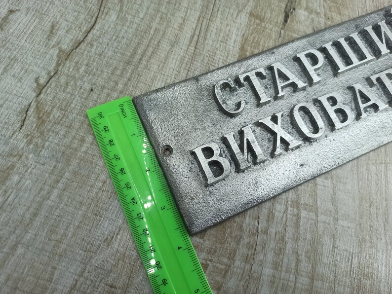 Puede incluir: Un letrero de metal plateado con el texto "СТАРШИ ВИХОВАТ" en letras en relieve. El letrero mide aproximadamente 15 cm de largo y 5 cm de ancho.