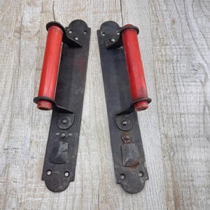 May include: A pair of vintage door handles. Each handle features a dark metal plate with a red, cylindrical grip. The plates have decorative details and mounting holes. The handles are set against a light wood grain background.