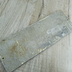 Puede incluir: Un letrero de metal rectangular desgastado con una superficie pintada de blanco descolorida. El letrero tiene un aspecto ligeramente oxidado y est&aacute; en blanco.