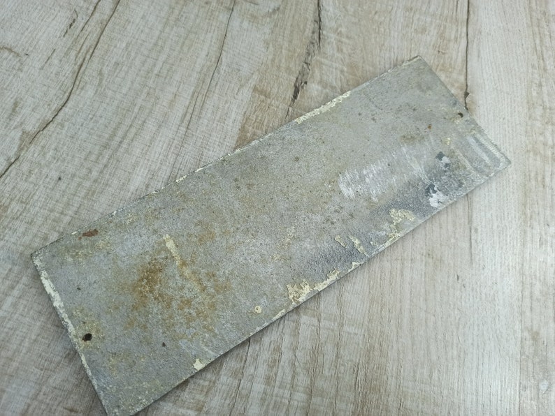 Puede incluir: Un letrero de metal rectangular desgastado con una superficie pintada de blanco descolorida. El letrero tiene un aspecto ligeramente oxidado y est&aacute; en blanco.