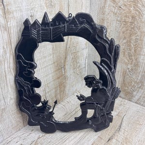 Puede incluir: Arte mural decorativo negro con una abertura circular central. El arte representa un castillo, una figura con arco y flecha y follaje estilizado. El marco tiene un acabado mate oscuro, agregando profundidad al diseño.