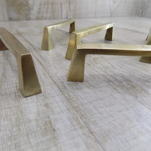 Op de afbeelding: Vijf messing kastgrepen met een geometrisch ontwerp. De grepen zijn in een rij op een licht houten oppervlak gerangschikt.