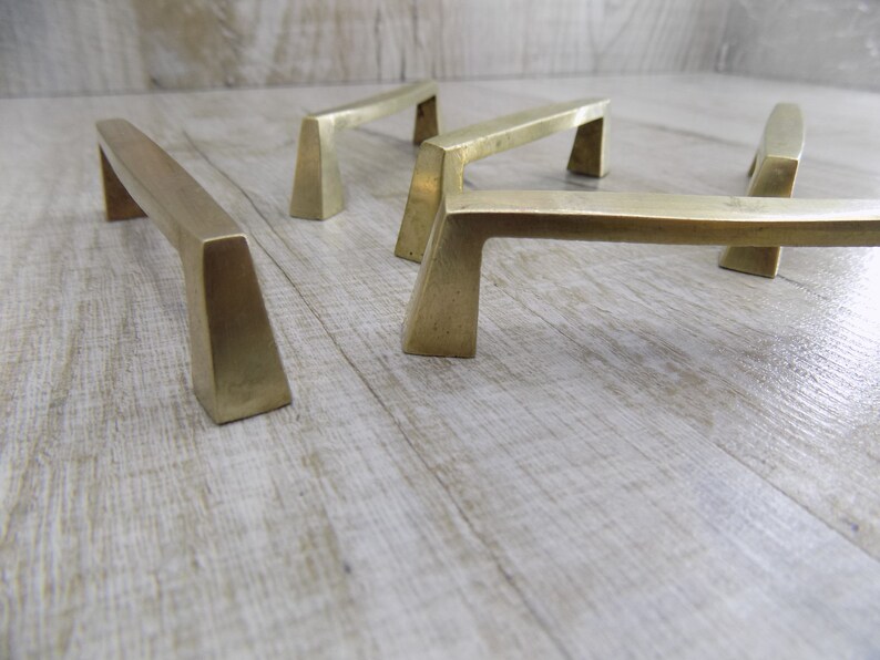 Op de afbeelding: Vijf messing kastgrepen met een geometrisch ontwerp. De grepen zijn in een rij op een licht houten oppervlak gerangschikt.