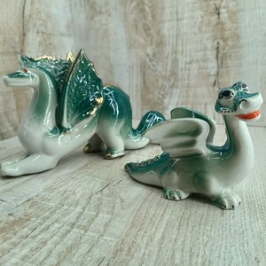 Peut inclure: Deux figurines de dragon en céramique, l'une plus grande que l'autre. Les deux sont vertes et blanches avec des accents dorés. Le plus grand dragon a des ailes et est en position accroupie. Le plus petit dragon est debout, les ailes déployées.