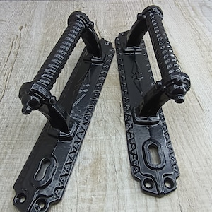 May include: Pair of black cast iron door handles with a decorative, twisted design. Each handle has a keyhole and a rectangular backplate with a patterned edge.