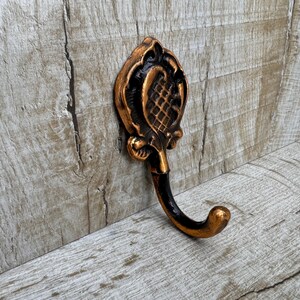 May include: A decorative metal hook with an ornate design in black and gold. The hook is mounted on a wooden surface.