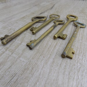 May include: A set of six antique brass keys with varying sizes and designs. The keys are arranged in a row on a light brown wooden surface.
