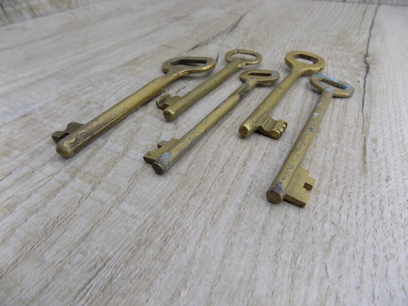 May include: A set of six antique brass keys with varying sizes and designs. The keys are arranged in a row on a light brown wooden surface.