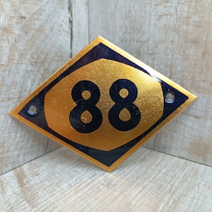 Könnte beinhalten: Ein goldfarbenes, rautenförmiges Hausnummernschild mit den schwarzen Zahlen "88" auf blauem Hintergrund.