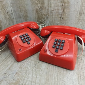 May include: Two red toy rotary dial telephones with black buttons and a white cord. The phones have the numbers 1-9 and 0 on the dial.