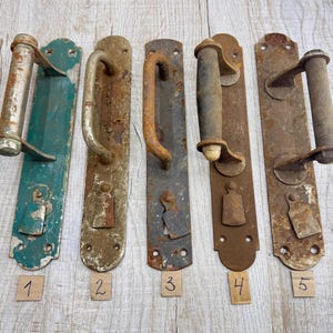 May include: Five antique door handles with varying designs and levels of rust. The handles are made of metal and are mounted on rectangular plates. One is painted teal, and the others are various shades of brown.