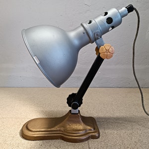 May include: A silver and black vintage desk lamp with a gold base and a flexible arm. The lamp has a large, round shade and a cord with a black and white braided pattern.