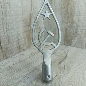May include: A silver metal finial with a star and hammer and sickle design. The finial is shaped like a teardrop and has a round base.