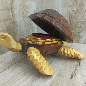Pode incluir: Uma caixa de madeira para joias em forma de tartaruga com tampa articulada. A tartaruga é esculpida com detalhes e tem um acabamento castanho e castanho claro. A caixa está aberta e tem um interior de veludo vermelho.