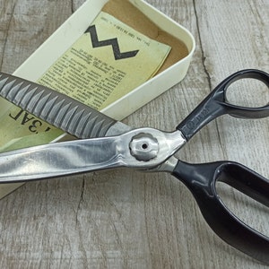 May include: A pair of black handled shears with a silver blade and a serrated edge. The shears are open and the blade is resting on a white box with text on it. The text on the box reads "3A1."