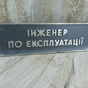 Op de afbeelding: Een grijs metalen bord met zilveren letters met de tekst "Инженер по эксплуатации".