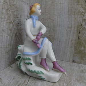 Puede incluir: Una figurita de porcelana de una mujer con un vestido blanco, una bufanda azul y patines de hielo rosas. Está sentada en una roca con hojas verdes y sostiene un par de guantes rosas.