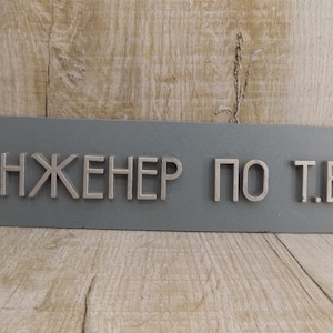 Op de afbeelding: Een grijs metalen bord met de tekst "ИНЖЕНЕР ПО Т.Б." in zilveren reliëfletters.