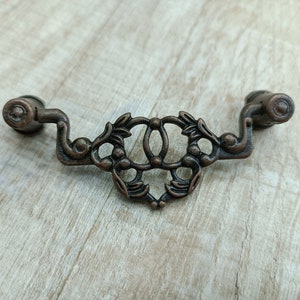 Op de afbeelding: Een decoratieve, sierlijke, bronzen kleurige ladegreep met een bloemmotief.