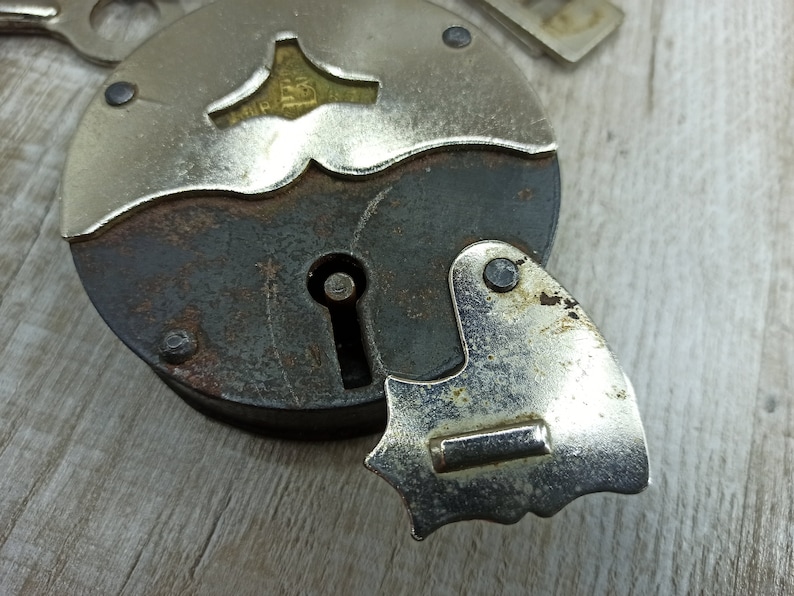 May include: A vintage silver padlock with a keyhole and a decorative design on the top. The padlock has a rusty brown finish and a silver metal latch.