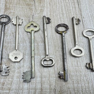 May include: Collection of vintage metal keys in various shapes and sizes. The keys are silver and gold-toned, with intricate designs on the key heads and teeth. The keys are arranged on a light-colored wooden surface.