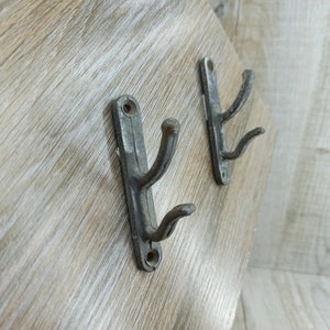 May include: Two antique metal hooks with a rustic finish. The hooks are mounted on a wooden surface and have a double prong design.