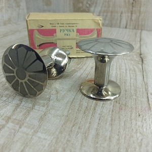 May include: Two silver metal cabinet knobs with a flower design. The knobs are on a wooden surface. The text on the box reads "РУЧКА РК1".