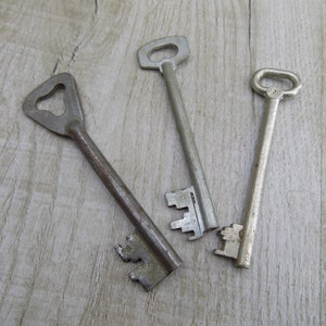 May include: Three antique keys with different designs and finishes, lying on a light wood surface.