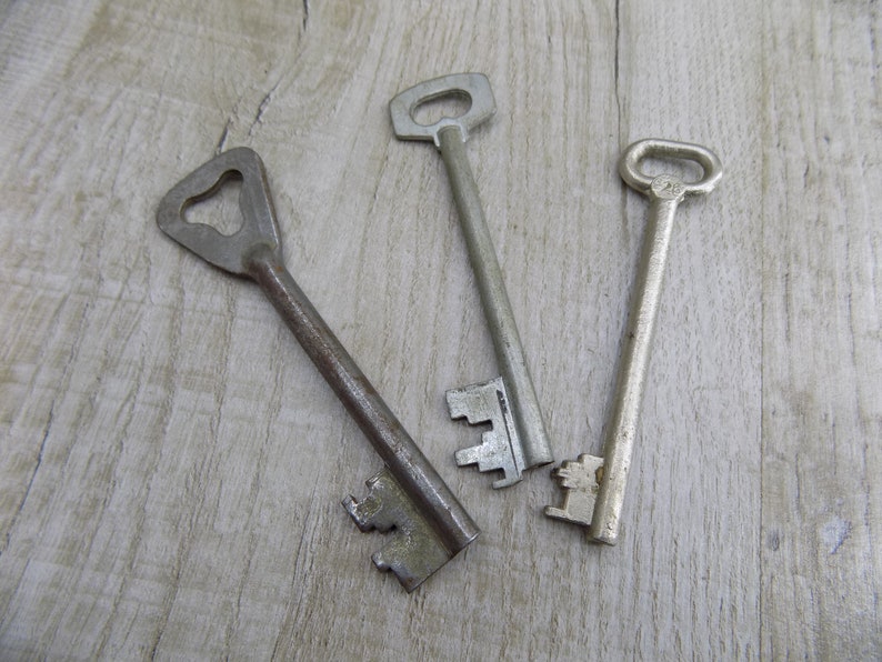 May include: Three antique keys with different designs and finishes, lying on a light wood surface.
