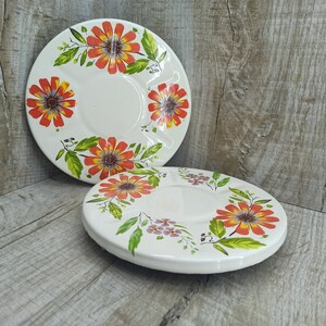 May include: Two white ceramic plates with a floral design. The plates have a raised rim and a center well. The flowers are orange, red, and yellow with green leaves.
