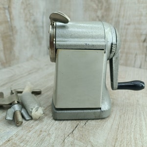Op de afbeelding: Een vintage zilveren potloodpuntenslijper met een zwarte draaiknop. De puntenslijper heeft een metalen basis en een transparant kunststof venster om het potlood te zien terwijl het wordt geslepen.