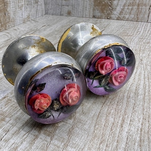 May include: Two round metal door knobs featuring a clear, domed glass insert. Inside each glass dome is a purple background with two three-dimensional pink rose blossoms and dark green leaves. The metal bases show signs of wear and gold-toned finish.