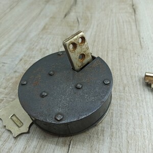 May include: A vintage, round, black metal lock with a silver keyhole and a silver key inserted into the lock. The lock is sitting on a light brown wooden surface.