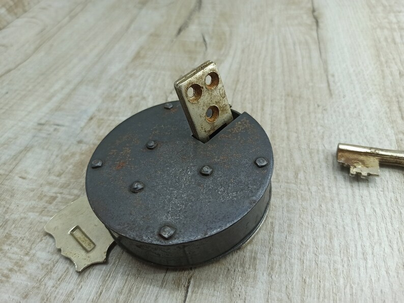 May include: A vintage, round, black metal lock with a silver keyhole and a silver key inserted into the lock. The lock is sitting on a light brown wooden surface.