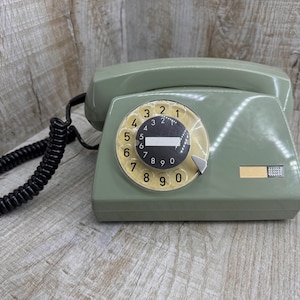 May include: A vintage green rotary dial phone with a black coiled cord. The phone has a round, cream-colored dial with black numbers 1 through 9 and 0.