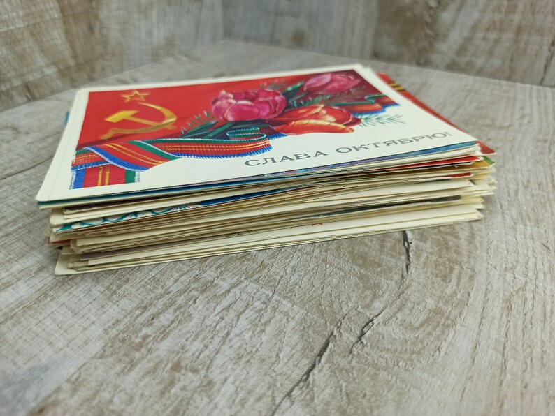 May include: A stack of vintage postcards featuring a red background with a gold star, hammer and sickle, and a bouquet of red tulips. The postcards have text in a foreign language.