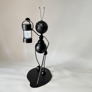 May include: A whimsical black metal ant figurine holding a small lantern with a glowing light. The ant has a smiling face and is standing on a black, oval-shaped base. The lantern is black with a clear, textured light.