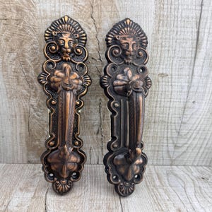 Puede incluir: Dos manijas de puerta ornamentadas de color bronce. Cada manija presenta una cabeza de león en la parte superior, intrincados diseños florales y un agarre vertical. Las manijas miden aproximadamente 30 cm de largo, con una estética antigua detallada.