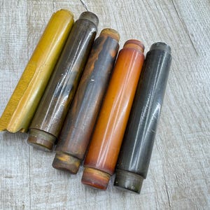 Puede incluir: Colección de seis objetos cilíndricos en tonos marrones, naranjas y amarillos. Los objetos tienen un acabado liso y brillante y parecen estar hechos de madera o un material similar. Están dispuestos en diagonal sobre una superficie de madera clara.