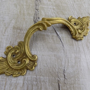 May include: A gold-toned ornate metal drawer pull with a decorative, scroll-like design.