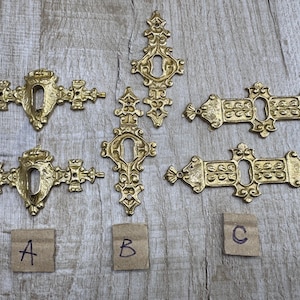 May include: A collection of five gold-toned metal keyhole escutcheons featuring ornate, symmetrical scrollwork designs. The pieces are arranged in three groups labeled with small cardboard squares marked with the handwritten letters A, B, and C.