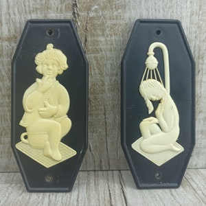 May include: Two black and white bathroom signs with a raised design. The sign on the left features a girl sitting on a toilet. The sign on the right features a girl showering.
