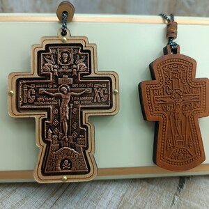 May include: Two wooden crosses with intricate carvings and religious iconography. The cross on the left has a copper-coloured finish and features a depiction of Jesus on the cross. The cross on the right is a natural wood colour and has a more stylized design.